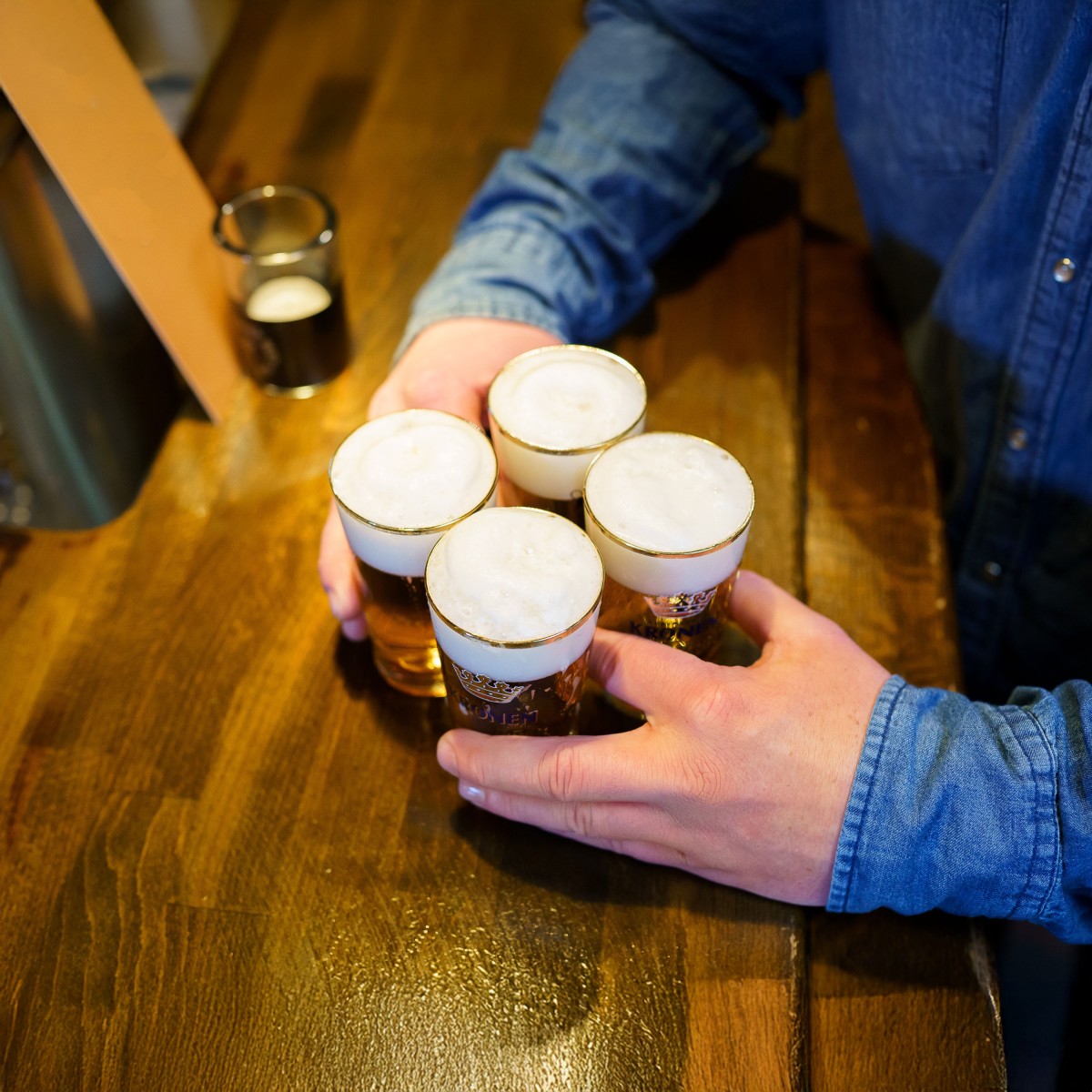 Eine Hand trägt vier Gläser Bier mit Schaum auf einer Holztheke.