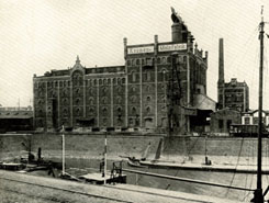 Historisches Industriegebäude mit Türmen und Schornstein am Flussufer, vorne liegt ein Boot.