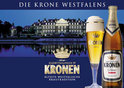 Ein großes Gebäude bei Nacht spiegelt sich in einem Gewässer, daneben steht ein Bierglas und eine Flasche.