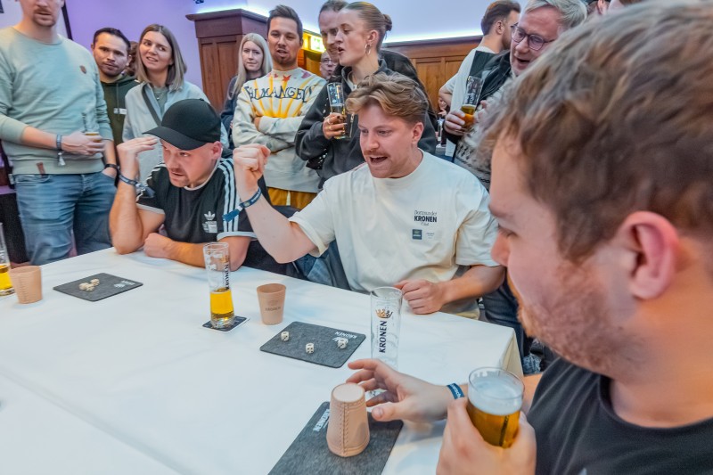 Drei Männer sitzen mit jeweils einem Bier an einem Tisch und spielen ein Würfelspiel. Um sie herum stehen mehrere Personen und sehen ihnen zu.