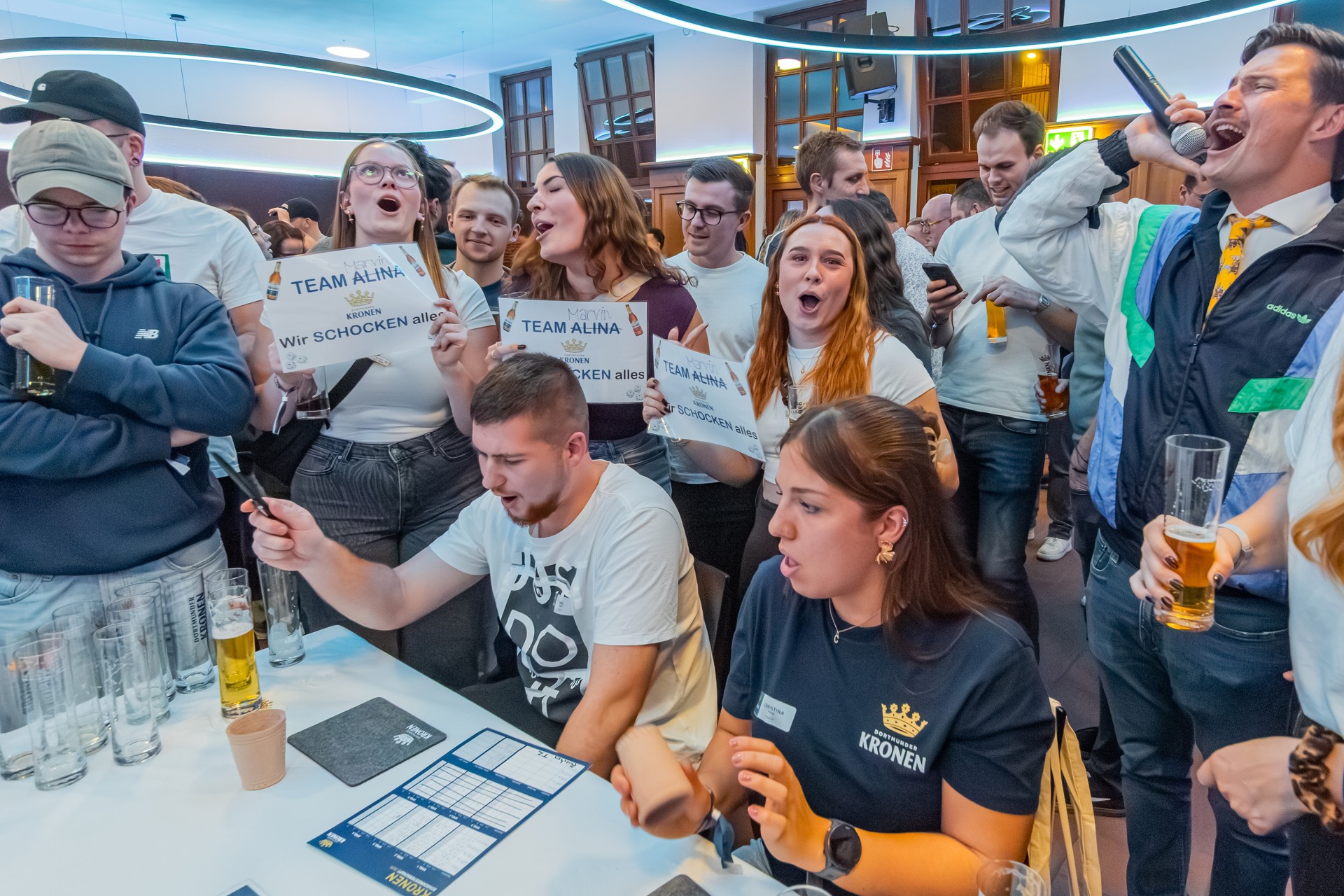 Zwei Männer sitzen an einem Tisch und spielen ein Würfelspiel. Um sie herum stehen mehrere Personen und verfolgen das Spiel.
