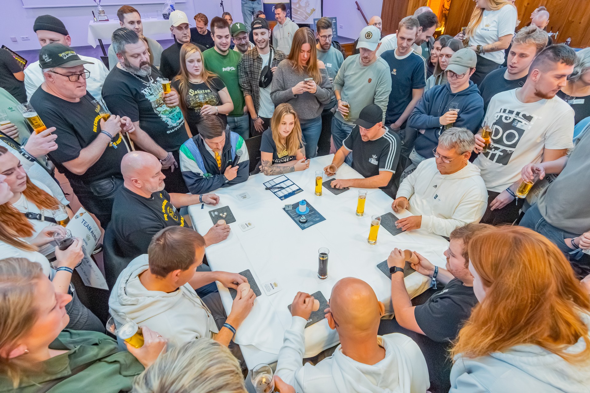 Mehrere Personen sitzen an einem Tisch und spielen ein Würfelspiel. Um sie herum stehen im Kreis mehrere Personen und beobachten den Spielverlauf.