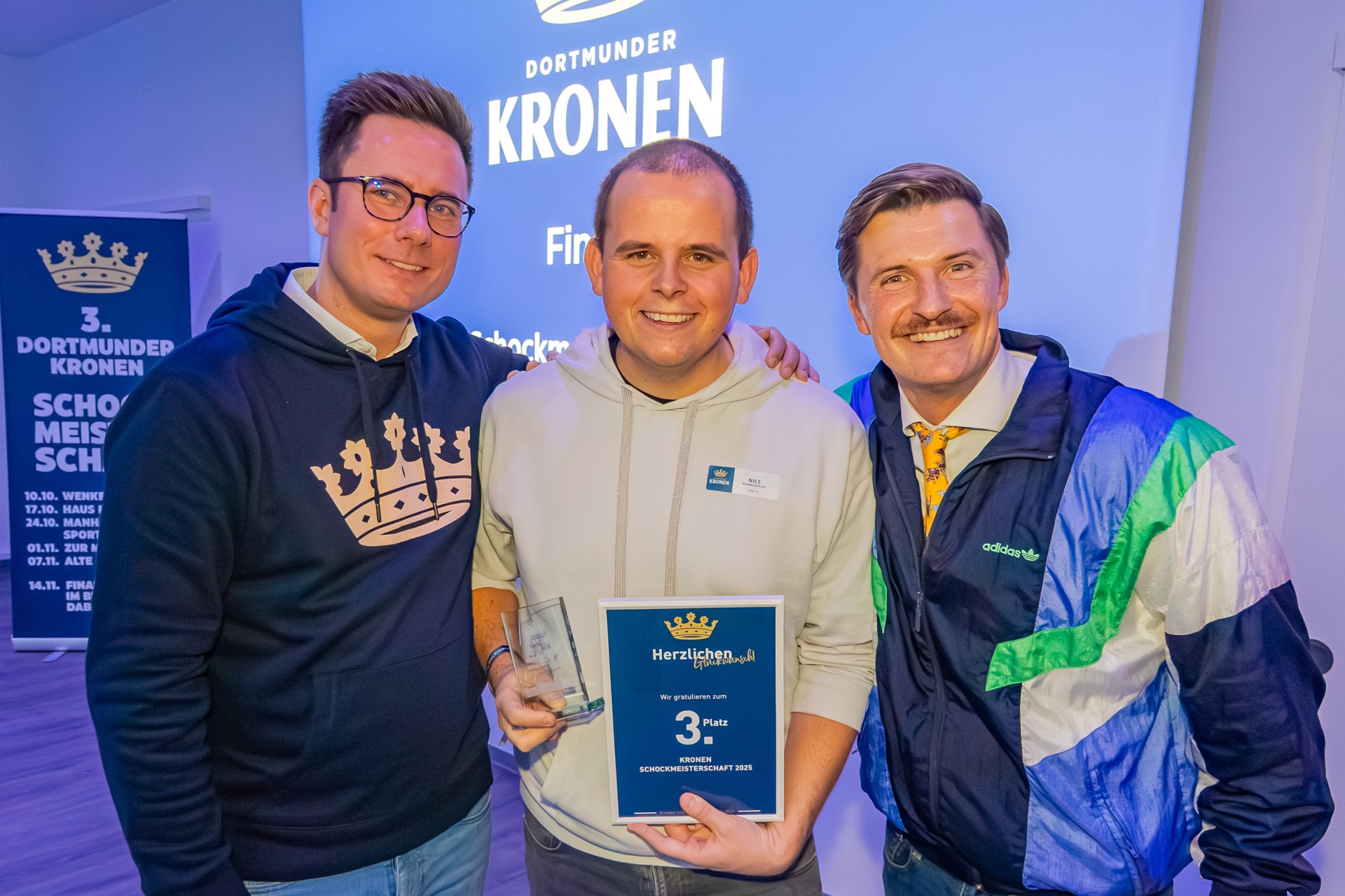 Drei Männer stehen nebeneinander. Der Mann in der Mitte hält eine Glastrophäe und eine Urkunde mit den Worten 