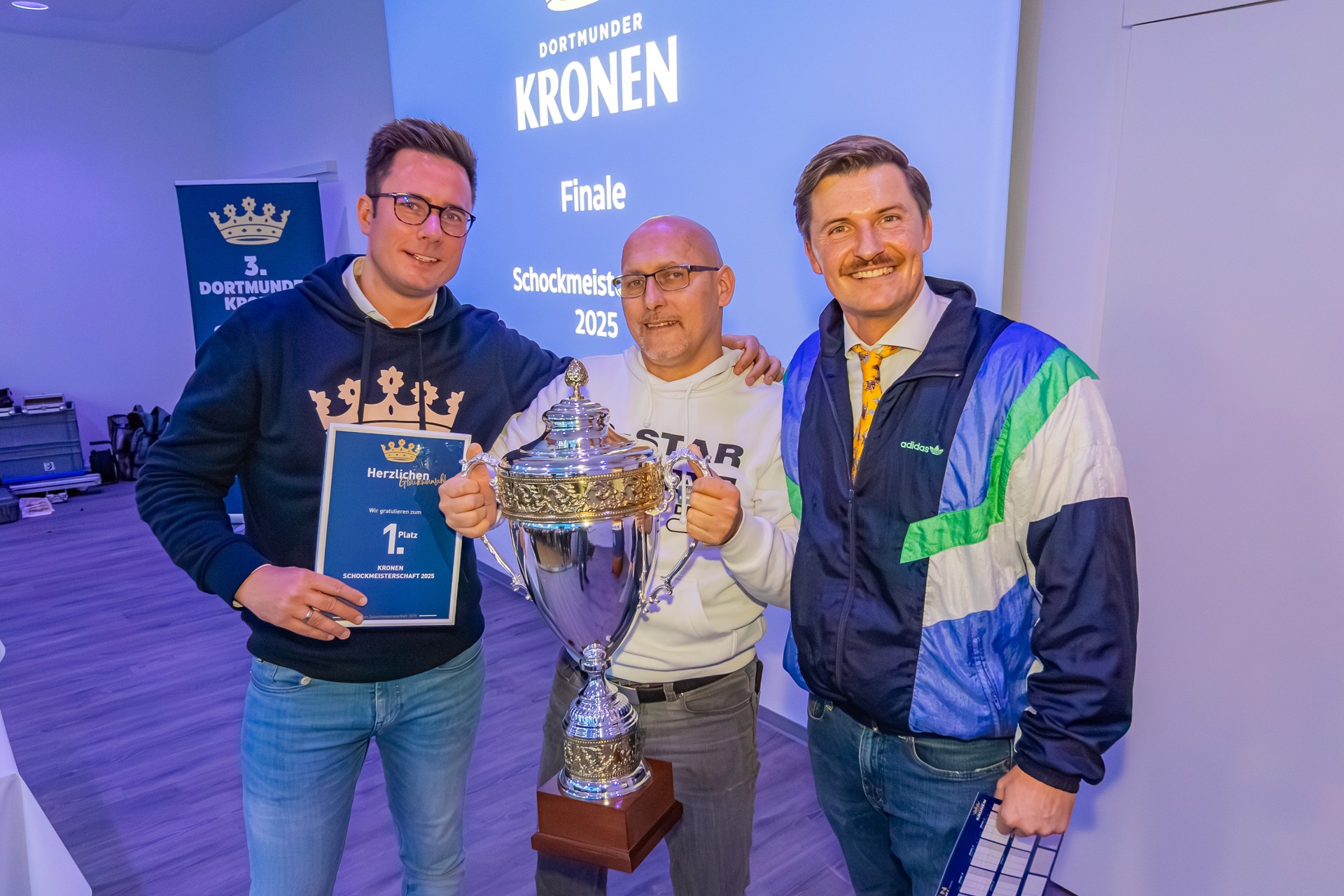 Drei Männer stehen nebeneinander. Der Mann in der Mitte hält eine Trophäe hoch. Der Mann links hält eine Urkunde mit den Worten 