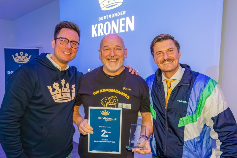 Drei Männer stehen nebeneinander. Der Mann in der Mitte hält eine Glastrophäe und eine Urkunde mit den Worten 