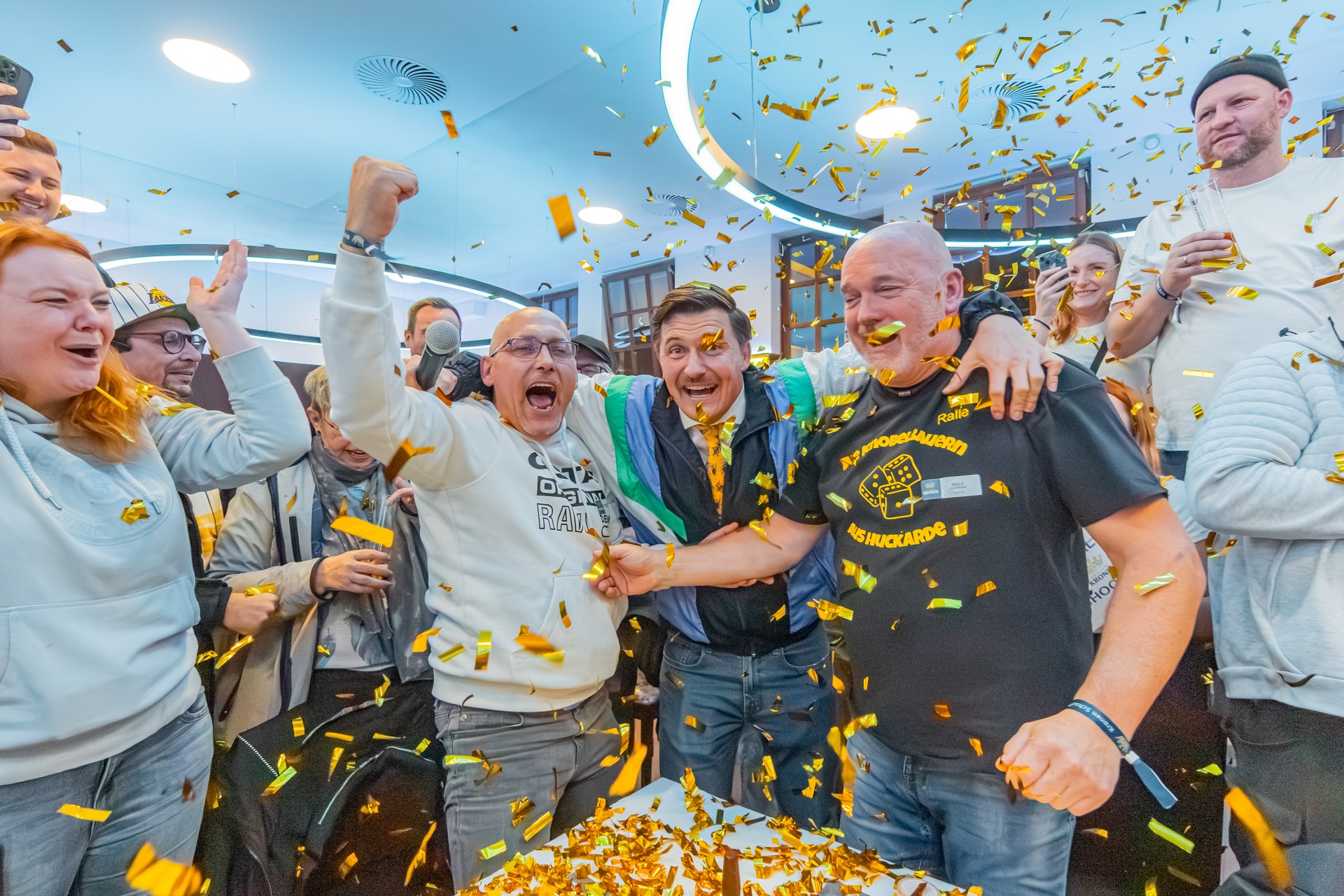 Drei Männer jubeln. Von der Decke fällt goldenes Konfetti. Im Hintergrund stehen weitere Menschen, die sich freuen.