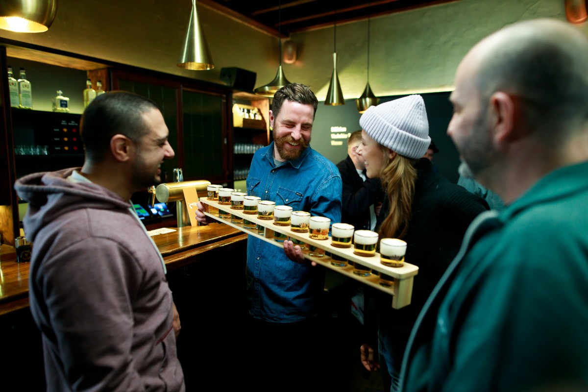 Ein Mann serviert lachenden Gästen in einer Bar kleine Biere auf einem Holzbrett. Sie stehen am Tresen und unterhalten sich angeregt.