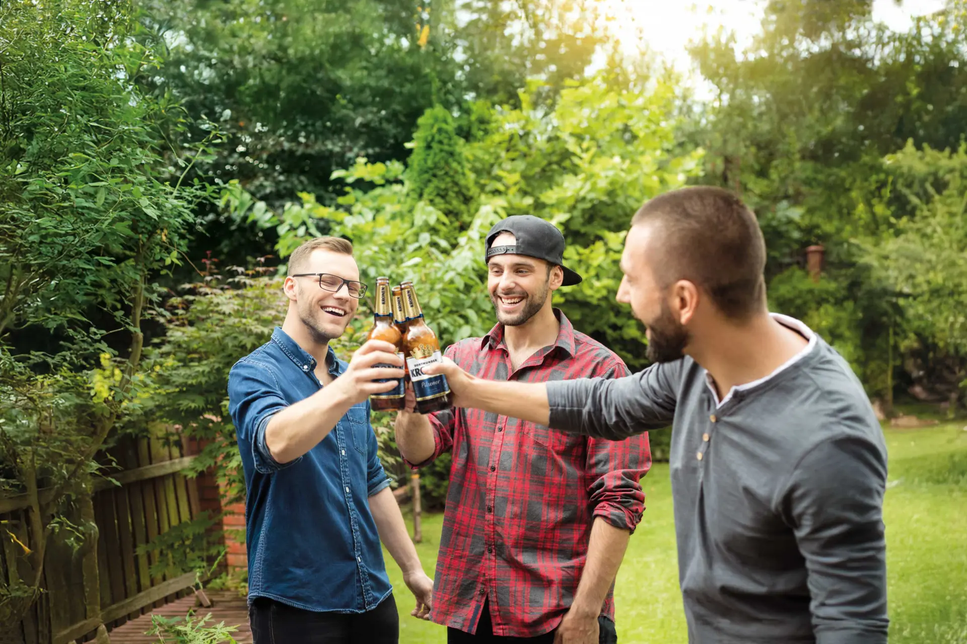 Drei Männer stoßen mit Bierflaschen an und lachen. Im HIntergrund grüner Garten mit Bäumen und Zaun.