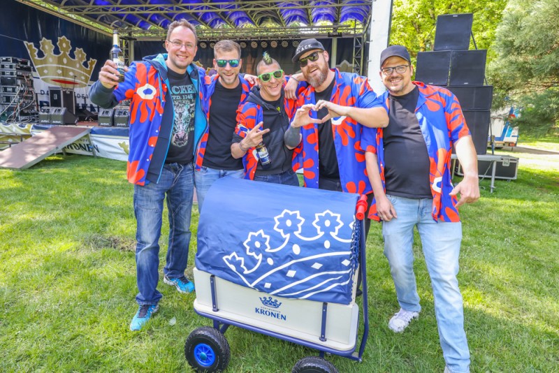 Vier Männer in farbenfrohen Jacken stehen lächelnd vor einer Bühne im Park. Sie halten Getränke und posieren mit einem kleinen Wagen.