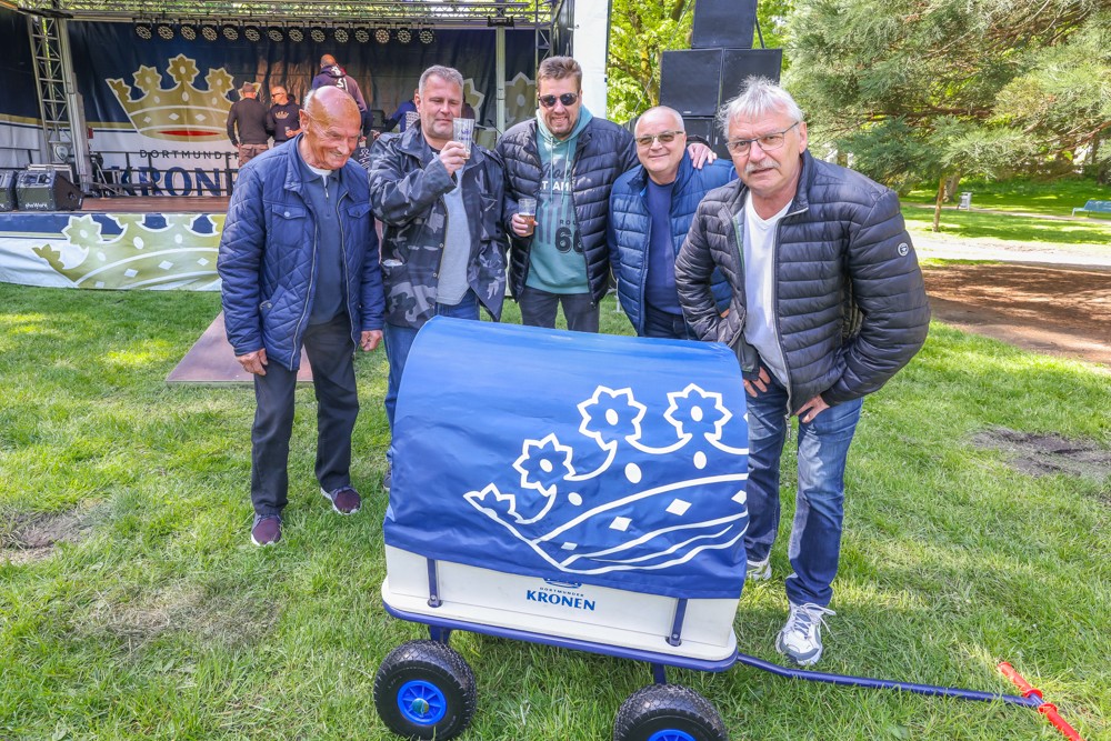 Fünf Männer stehen im Freien bei einem Festival neben einem blauen Handwagen.