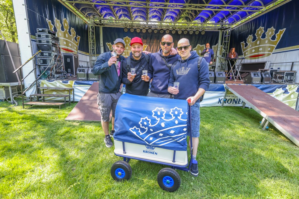 Vier Männer stehen lächelnd vor einer Festivalbühne und halten Getränke, während einer einen Bollerwagen festhält.