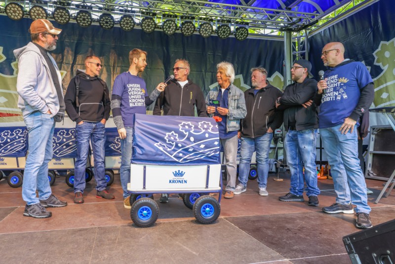 Acht Männer stehen neben einem Wagen mit blauen Rädern auf einer Bühne.