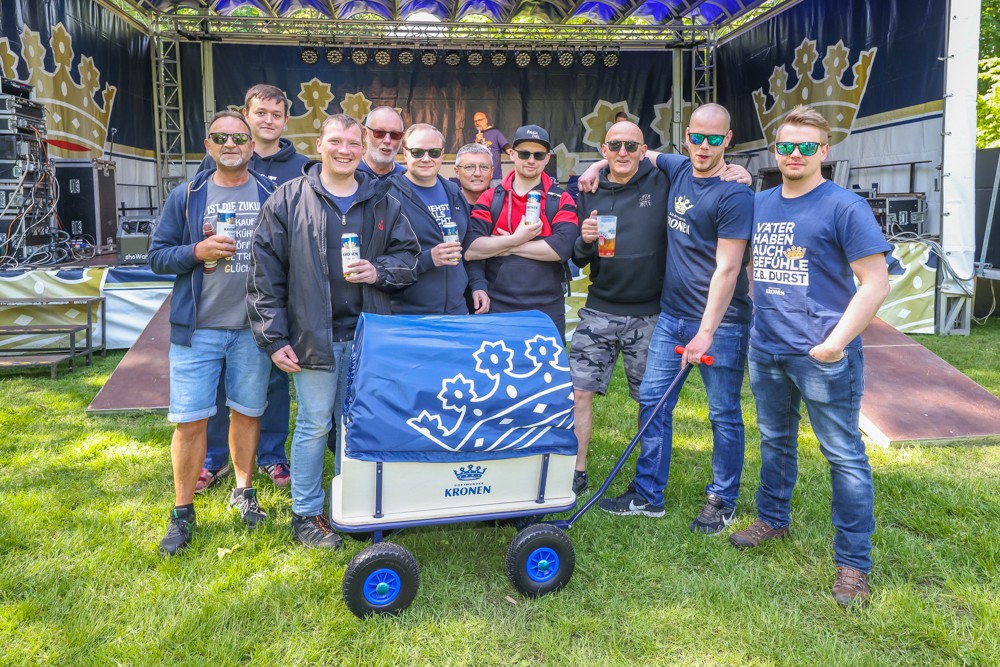 Zehn Männer stehen mit Getränken im Freien vor einer Bühne, neben einem Wagen.