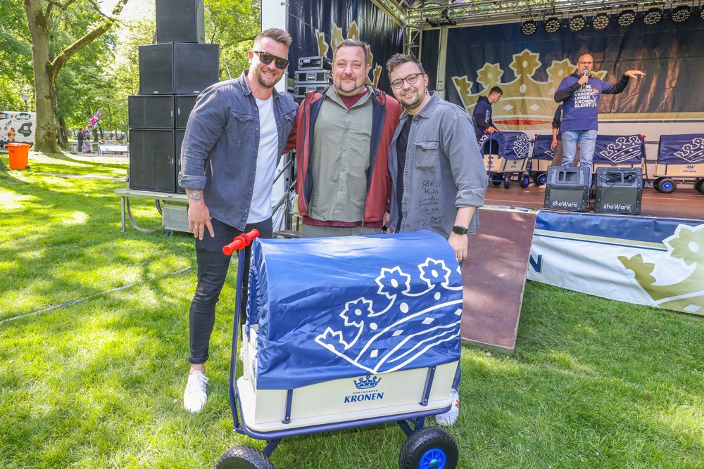Drei Männer stehen lächelnd neben einem blau-weißen Wagen auf einer Bühne im Park.