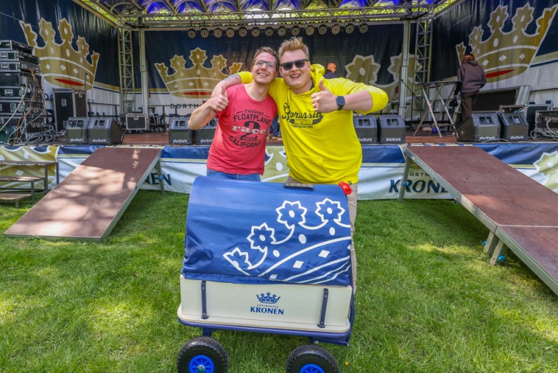 Zwei junge Männer stehen lächelnd neben einem kleinen blauen Wagen auf einer Wiese vor einer Bühne.