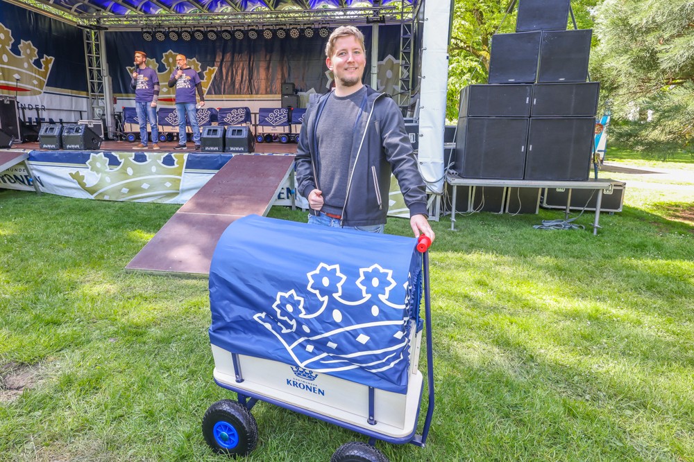 Ein Mann steht lächelnd auf einer Wiese und hält einen blauen Handwagen neben einer Bühne mit zwei Sängern.