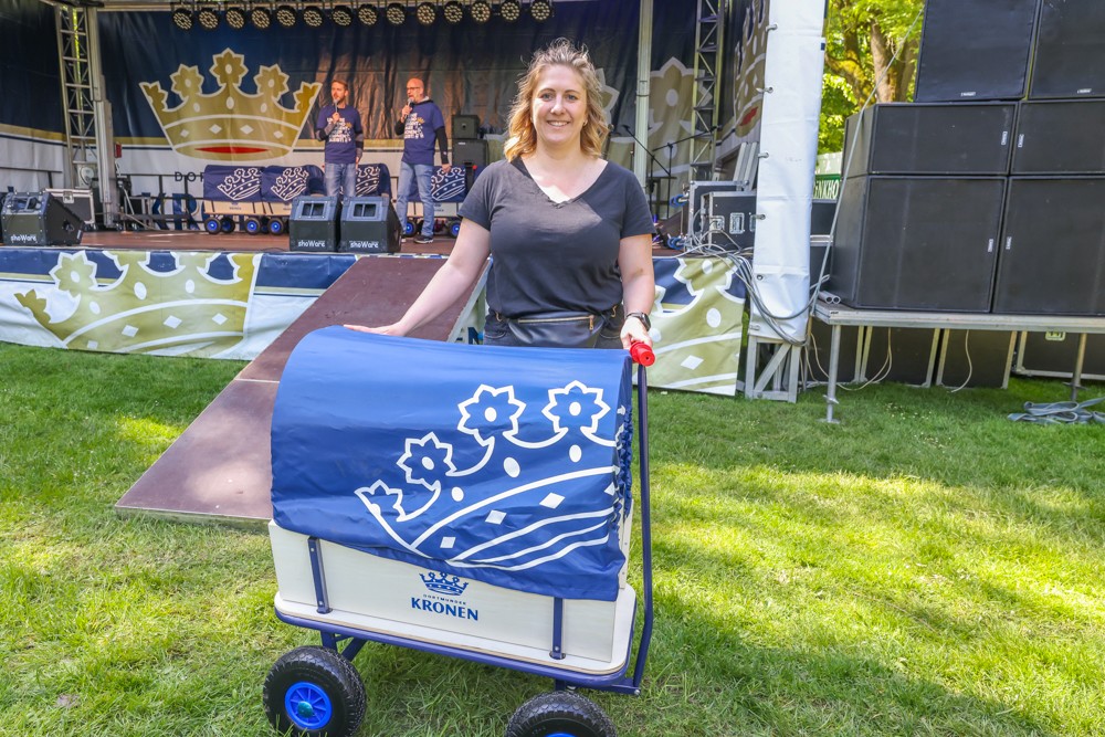 Eine Frau steht auf einer grünen Wiese und hält einen dekorierten Handwagen neben einer Bühne.