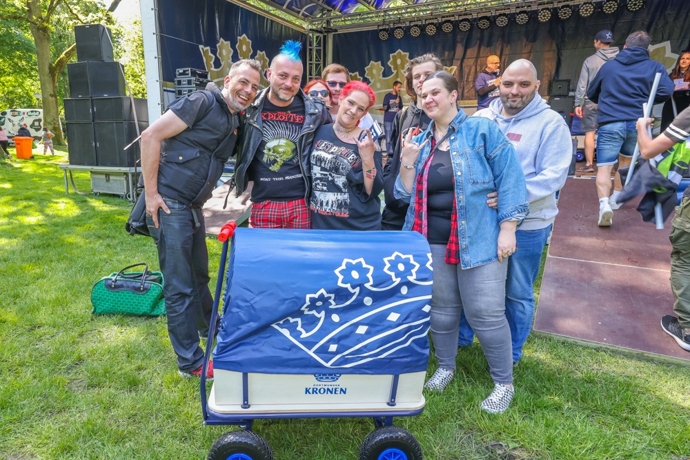 Sechs Personen stehen lächelnd vor einer Bühne im Park neben einem blauen Wagen.
