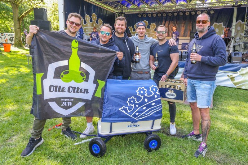 Sechs Männer stehen draußen mit Bierflaschen, einer hält eine Flagge und ein anderer einen Karren.
