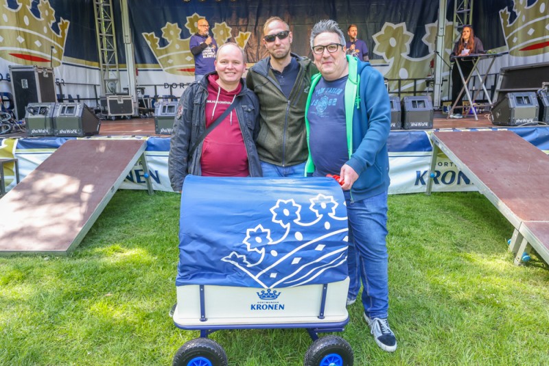 Drei Personen stehen lächelnd vor einer Bühne und halten einen blauen Handwagen.