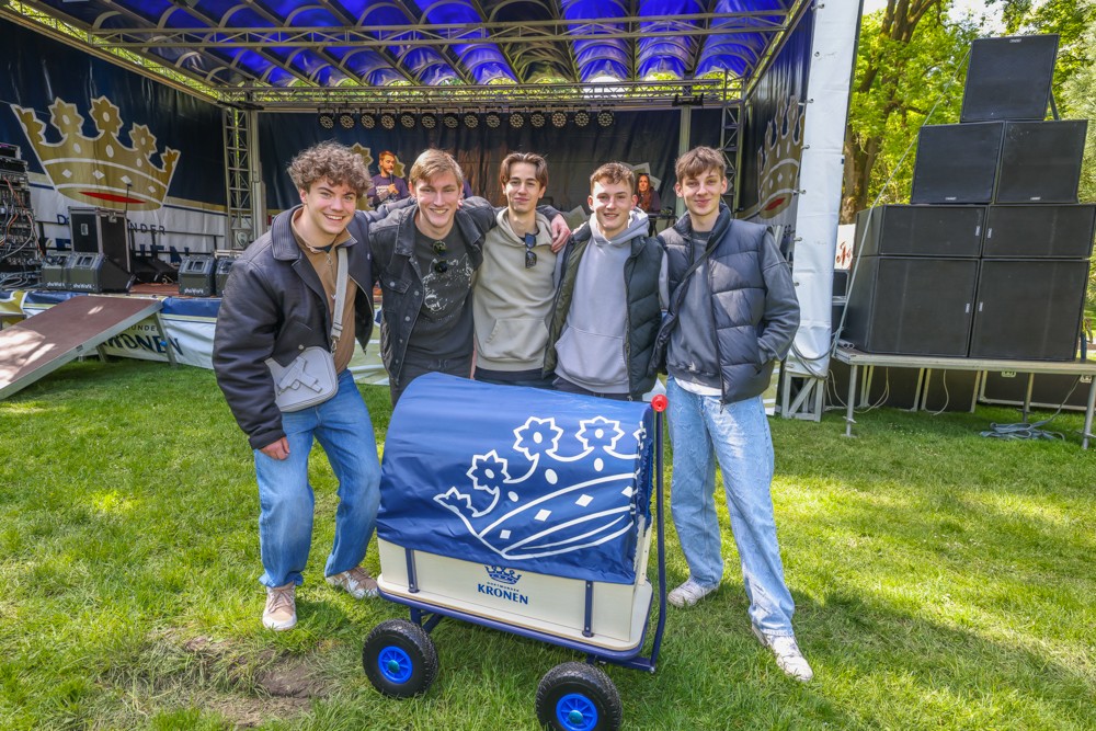 Fünf junge Männer stehen lächelnd vor einer Bühne mit einem blauen Bollerwagen.