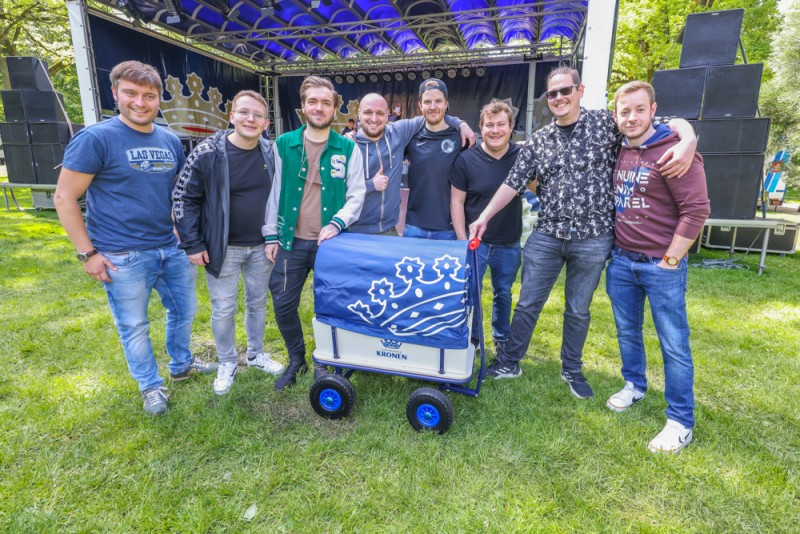 Acht Männer stehen lächelnd vor einer Bühne im Park, einer hält einen blauen Wagen.