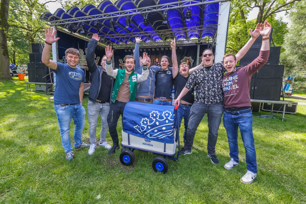 Sieben Männer jubeln fröhlich vor einer Bühne in einem Park, neben ihnen steht ein blauer Wagen.