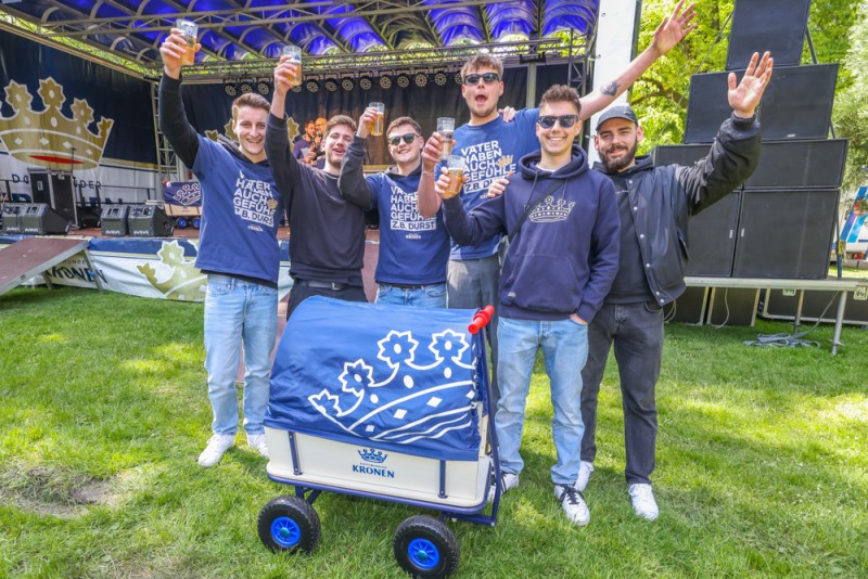 Fünf lächelnde junge Männer stehen im Freien und halten Getränke hoch.