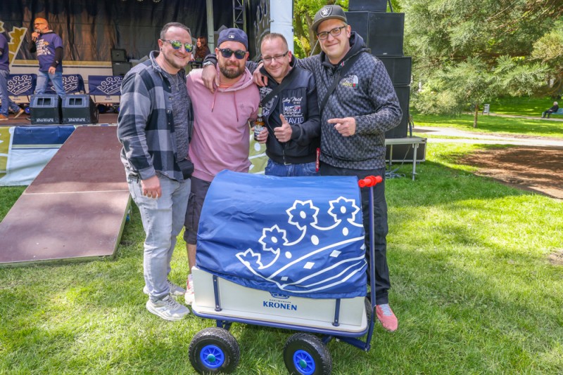 Vier Männer stehen lächelnd neben einem Bollerwagen im Park.