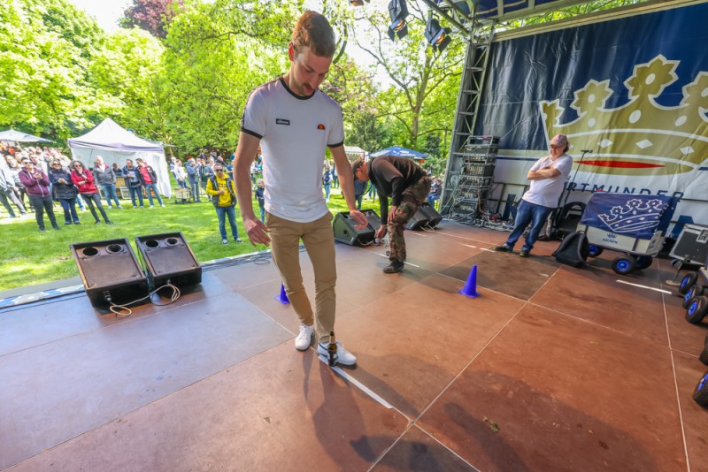 Ein junger Mann balanciert auf einem Rollbrett bei einem Event im Freien. Zuschauer beobachten seine Darbietung.
