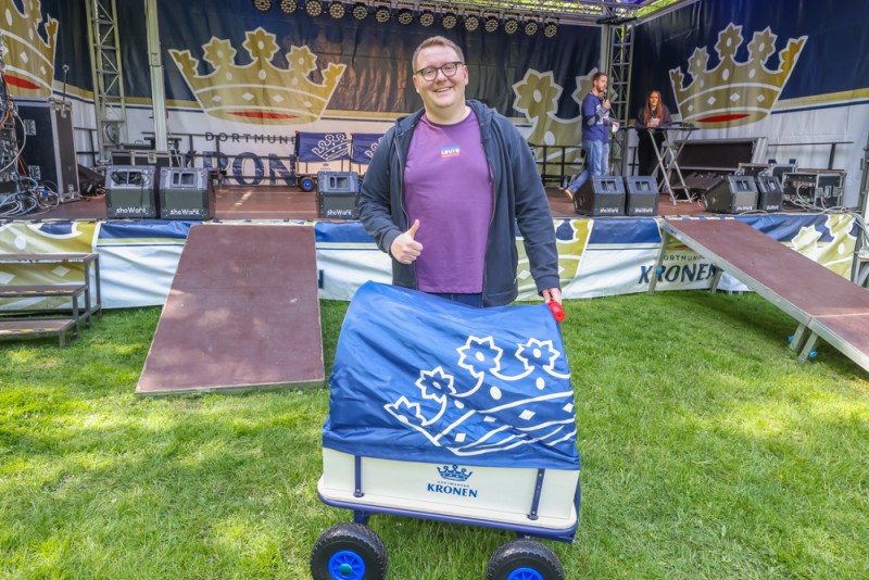 Ein Mann steht lächelnd vor einer Bühne und hält einen Wagen mit blauem Muster.