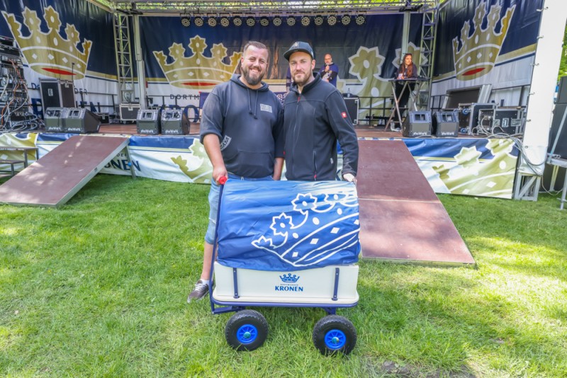Zwei Männer stehen neben einem Handwagen vor einer Bühne im Park.