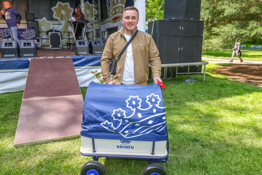 Ein Mann steht neben einem blauen Handwagen mit weißem Blumenmuster auf einer Wiese bei einem Bühnenereignis.