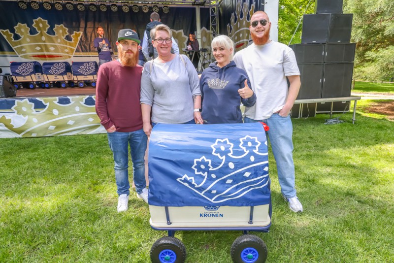Vier Personen posieren lächelnd neben einem blauen Wagen bei einem Festival.
