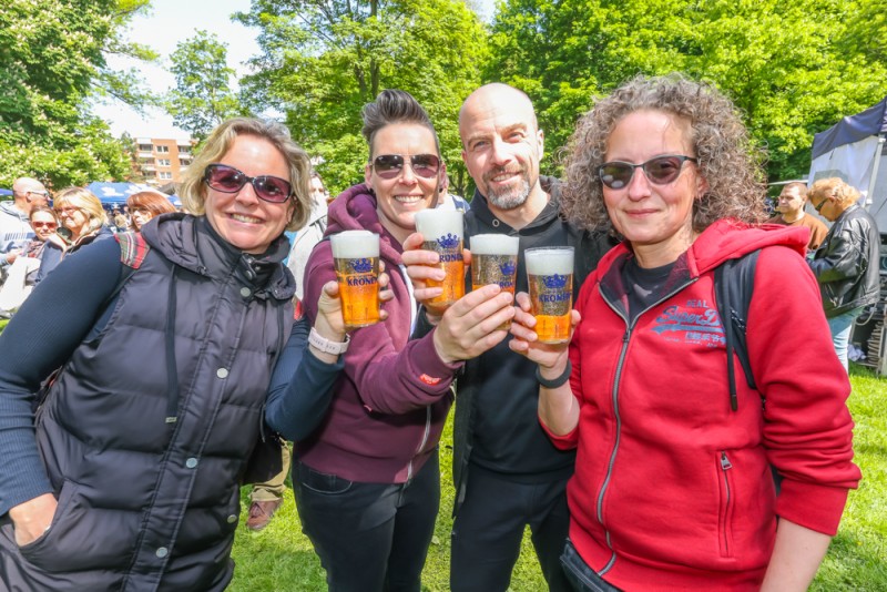 Zwei Frauen und zwei Männer lächeln in die Kamera und halten Biergläser in einer grünen Parkumgebung.