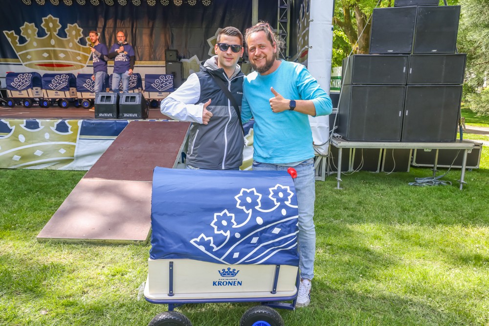 Zwei Männer stehen lächelnd neben einem geschmückten Wagen auf einer Veranstaltung im Freien.