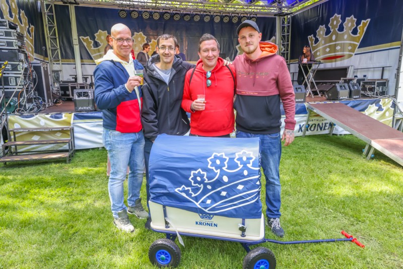 Vier Männer stehen lächelnd neben einem dekorierten Wagen auf einer Wiese.