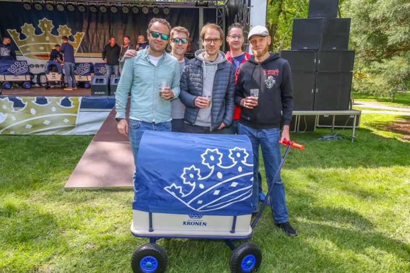Vier Männer stehen mit Getränken in einer Parkszene vor einer Bühne, neben ihnen ein Bollerwagen.