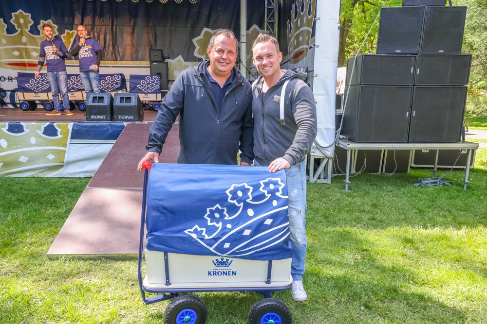 Zwei Männer stehen lächelnd neben einem Wagen mit blauer Flagge in einem Park.