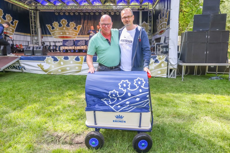 Zwei Männer stehen neben einem Wagen mit blauer Plane auf einer Wiese vor einer Bühne.