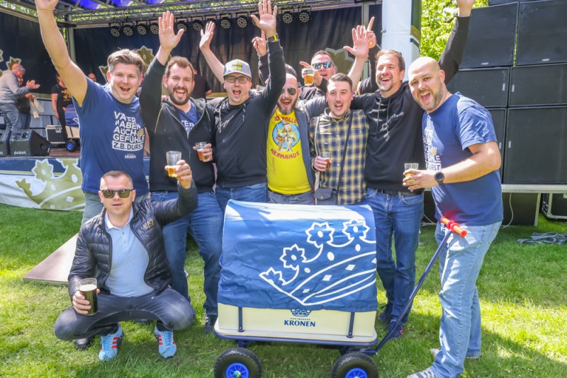 Gruppe fröhlicher Männer feiert im Freien bei einem Festival, einer hält einen Bollerwagen.