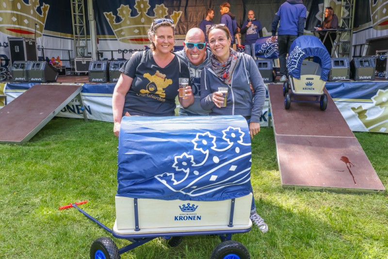 Drei Personen stehen lächelnd neben einem dekorierten Wagen bei einem Festival.