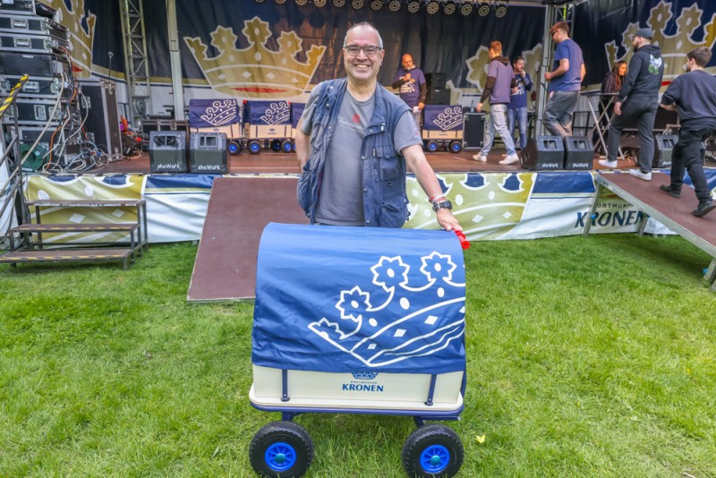 Ein Mann mit Brille steht lächelnd neben einem Bollerwagen auf einer Wiese. Hinter ihm ist eine Bühne mit Musikern zu sehen.