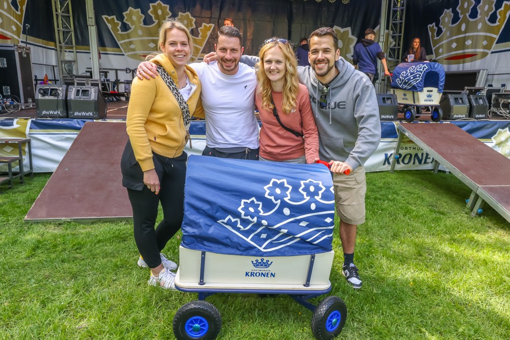 Zwei Frauen und zwei Männer stehen lachend neben einem Bollerwagen auf einer Wiese.