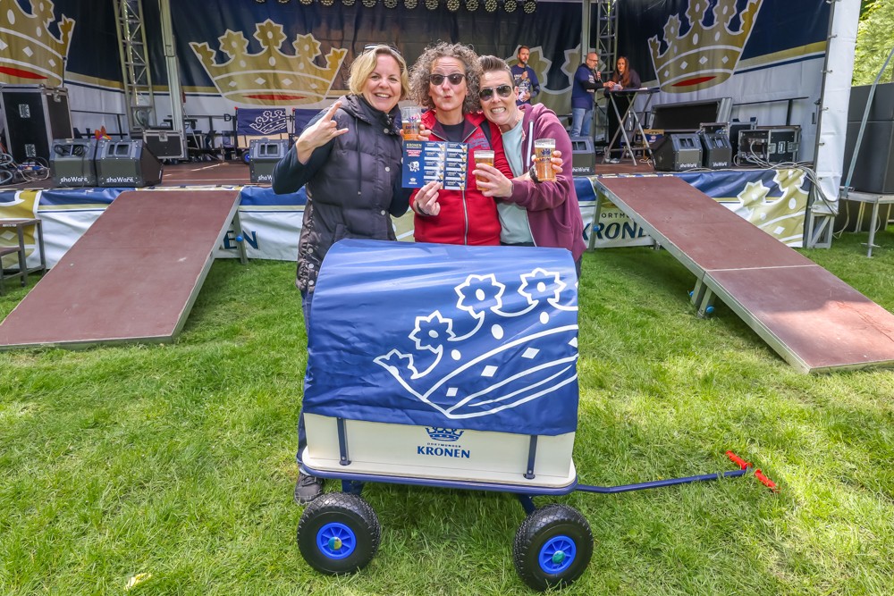 Drei Frauen stehen lächelnd vor einer Bühne und halten Getränke, neben ihnen steht ein blauer Handwagen.