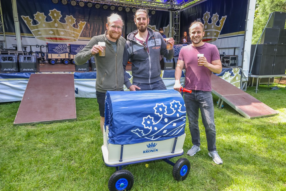 Drei Männer stehen im Freien, halten Gläser und lächeln neben einem dekorierten Wagen.