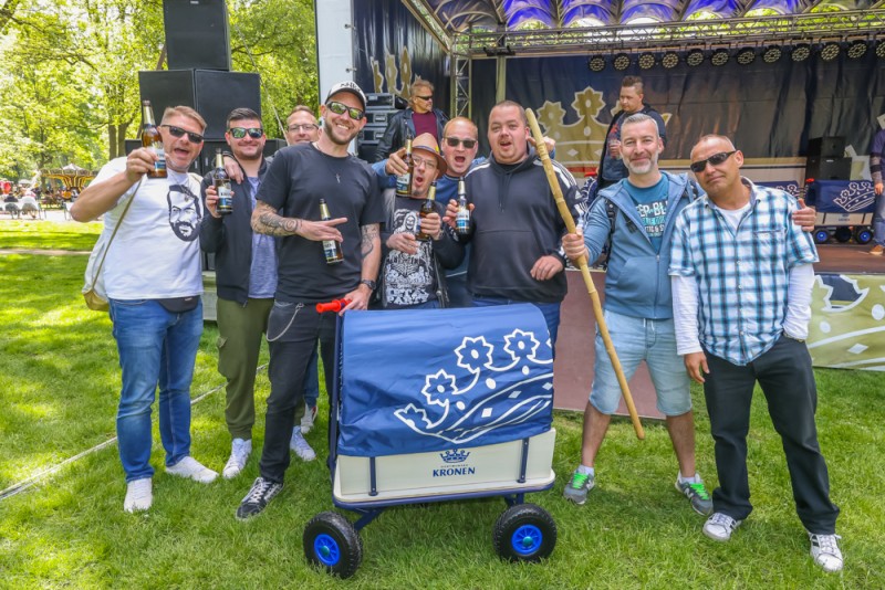 Acht Männer mit Getränken lächeln vor einer Bühne, neben einem blauen Bollerwagen.