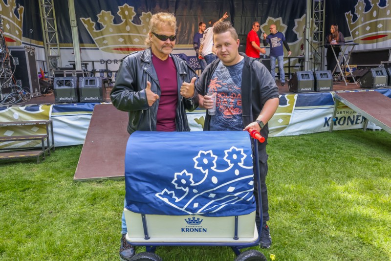 Zwei Männer stehen neben einem blauen Handwagen auf einer Wiese vor einer Bühne.