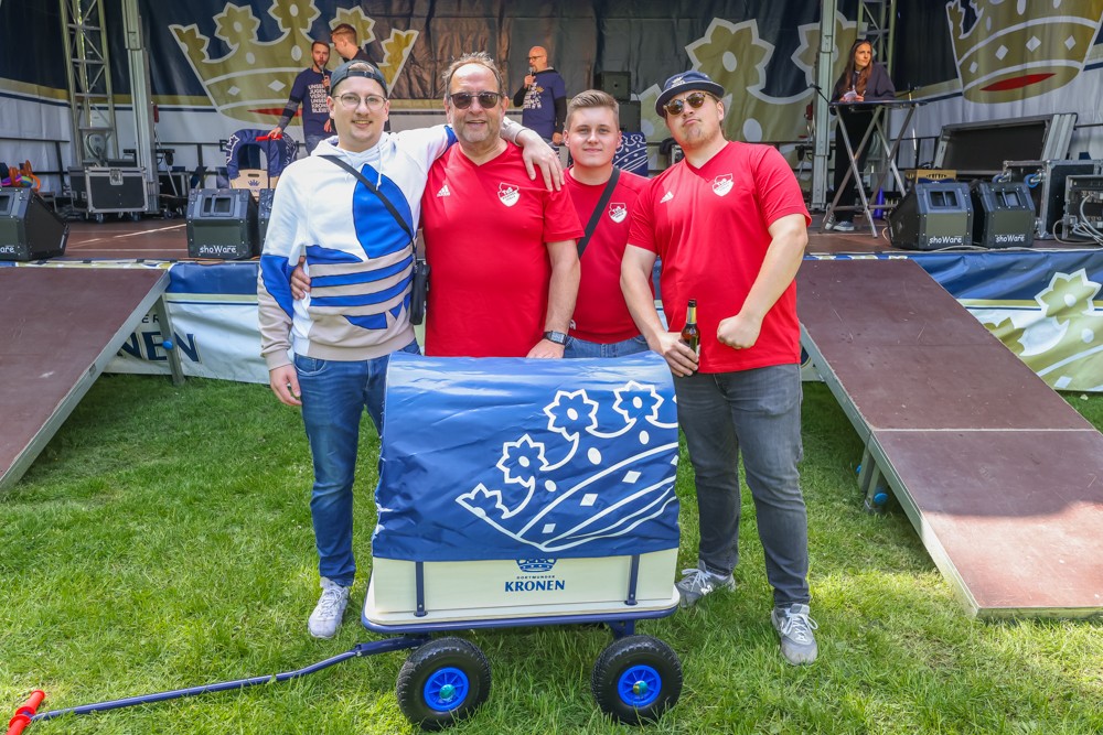 Vier Männer stehen vor einer Bühne neben einem blauen Wagen. Sie tragen lässige Kleidung und lächeln.