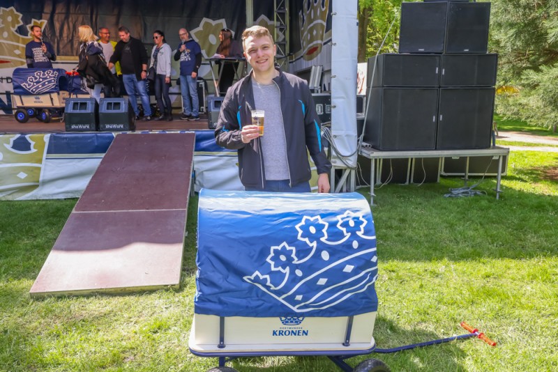 Ein junger Mann steht lächelnd mit einem Getränk vor einer Bühne im Park.